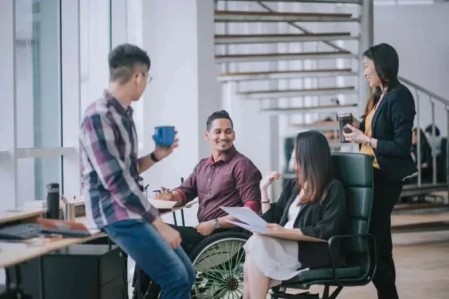 Indian white collar male worker in wheelchair having cheerful discussion leading conversation with colleague about ATS and CRM in an office.